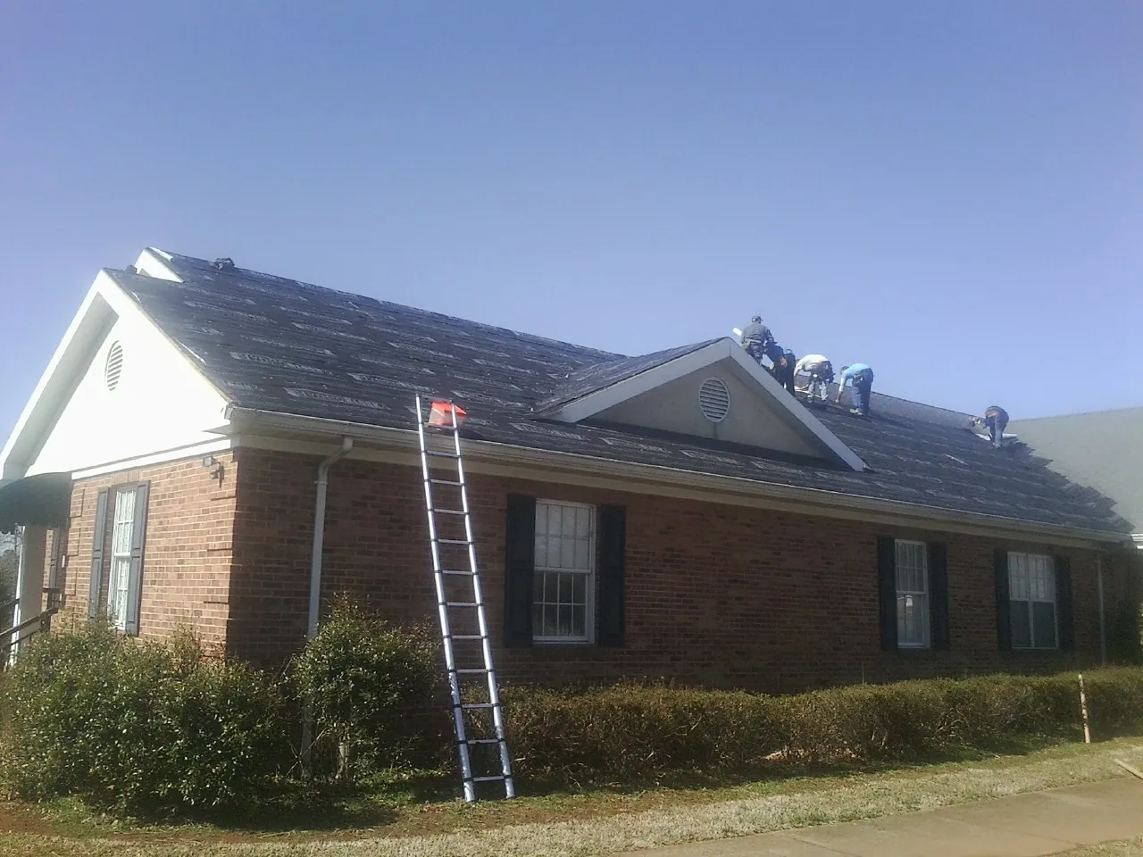 Crew tearing shingles off a brick ranch roof with a ladder set — Emergency Roof Repair work by our crew in Almont