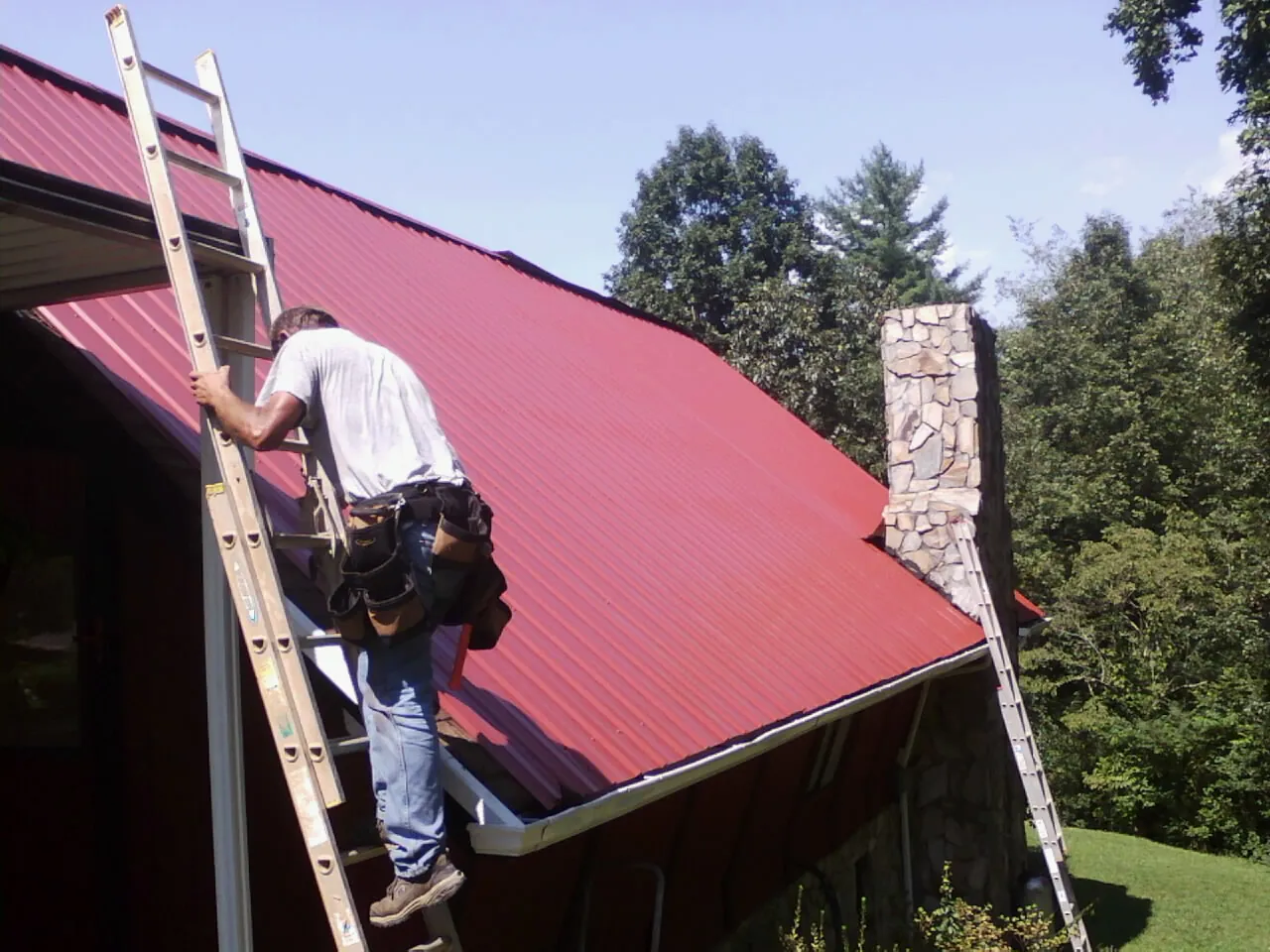 Roofer on a ladder beside a red metal roof and stone chimney — Soffit & Fascia Repair by Roofing Brothers in Almont